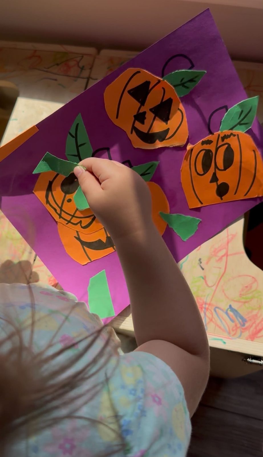 A child's hand gluing paper jack-o'-lanterns onto purple paper